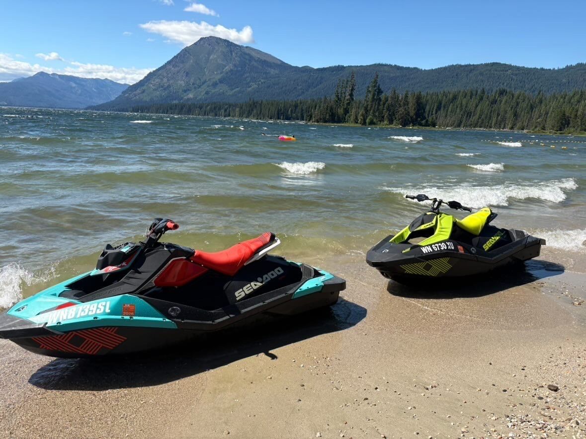 Two Sea-Doo Spark Trixx jet skis on a lakeshore with Cascade mountains behind